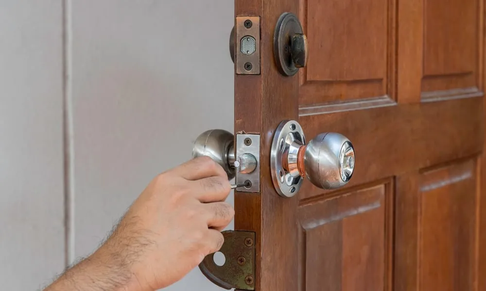 A close-up of a locksmith's hand using a tool to adjust or repair the latch mechanism on an open wooden door with silver doorknobs.