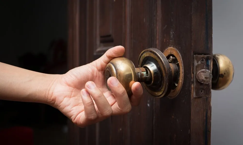 close-up of a person's hand turning a loose, old brass doorknob on a dark wooden door with exposed internal components.