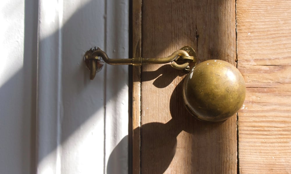 A close-up of an old wooden door featuring an antique brass doorknob and a traditional cabin hook latch secured to a white door frame.